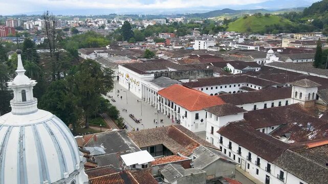 Drone panorama over Popayan central plaza showing a white domed church, long colonnade with red tile roofs, tree-lined park and pedestrians with colorful umbrellas.