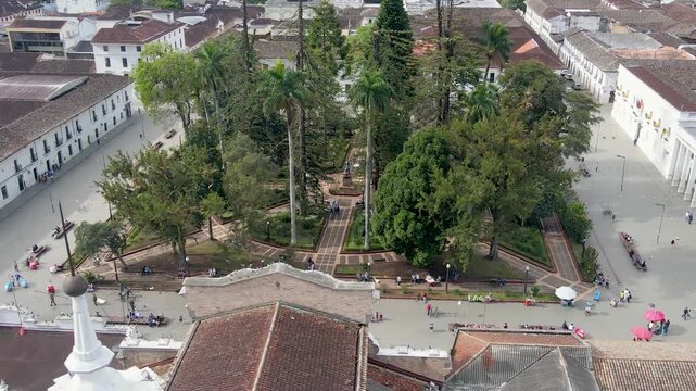 Drone reveal of a white cathedral dome over red-tiled roofs and a tree-filled central park, with winding paths, benches and small groups of people under colorful umbrellas.