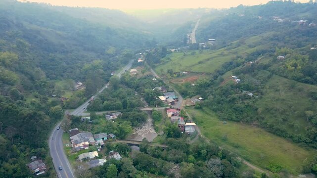 Drone aerial over green hills near Popayan showing a winding highway, small bridge crossing a river and a cluster of houses with colored roofs and light traffic.
