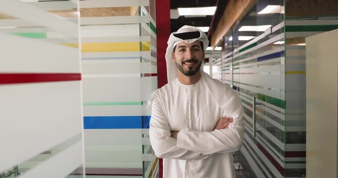 Confident young Middle Eastern business professional man in traditional Arabic dress standing in office corridor, keeping hands folded, looking at camera, getting happy, posing for head shot portrait