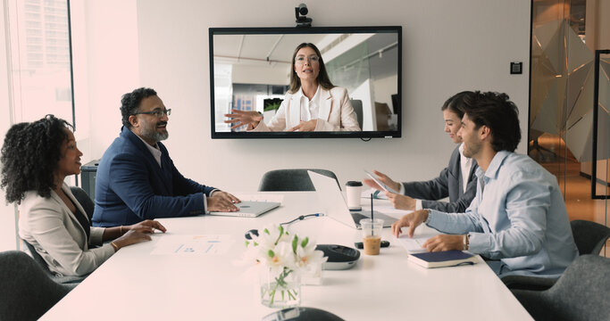 Diverse business team staff meeting at office table, talking to female company leader on online video call, using large TV display board for global communication, discussing teamwork