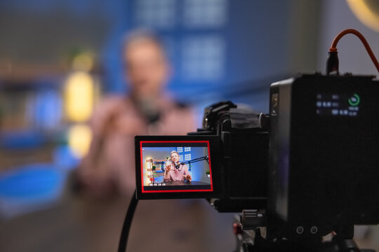 Closeup of video camera filming podcast session, monitor camcorder screen showing host influencer speaking during live broadcast stream production, blurred background. Digital media recording concept