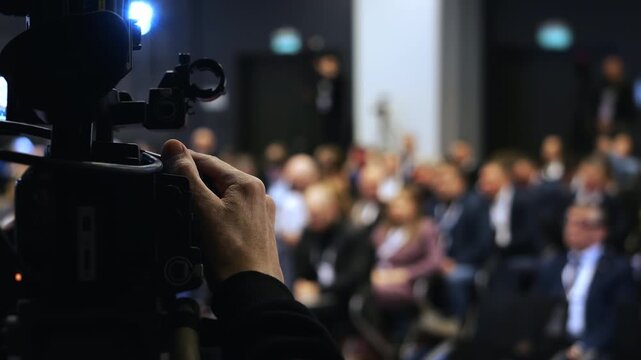 Camera operator filming audience at business conference or seminar with professional video equipment in modern event hall.
