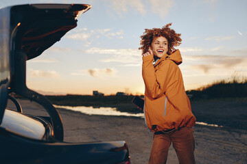 Fototapeta premium A happy woman in an orange jacket smiles outdoors near an open car trunk at sunset. The woman enjoys a casual lifestyle with natural curly hair and a relaxed pose.