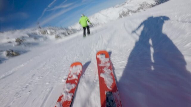 Skier in a green jacket skis down a snowy mountain. View from the perspective of the skier, showing their skis and shadow. Low angle slow motion