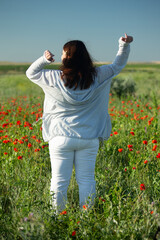Portrait of a happy young woman with her back against a backdrop of wildflowers, her arms raised. Body positivity. Vertical photo.