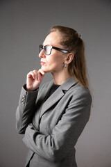 A profile portrait of a woman with glasses and long hair in a business suit, lost in thought and touching her chin. Vertical photo