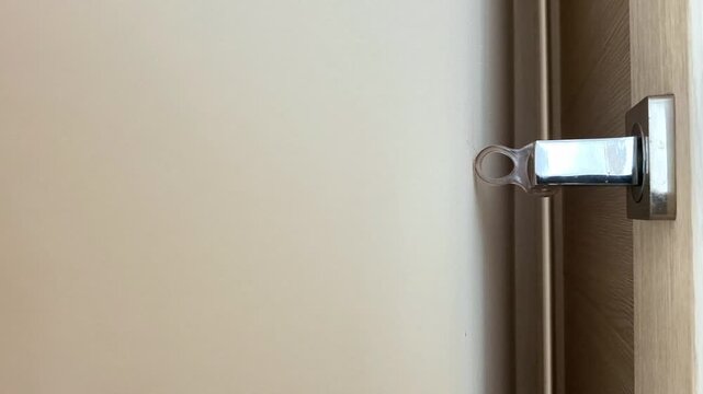 Close up of metallic door latch extending into strike plate on wooden frame, showing secure locking mechanism with beige wall background and detailed hardware view