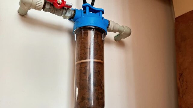 Wall mounted water filtration unit with transparent housing showing a brown sediment filled cartridge, connected to white PVC pipes, emphasizing plumbing and filtration maintenance