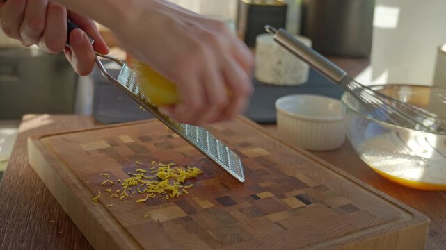 Close up of hands grating fresh lemon zest with metal grater on wooden cutting board in kitchen. Concept of cooking baking preparation and homemade food.