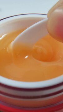 Close up of stirring thick orange liquid in red container with spoon, viscous skincare product isolated on white background.