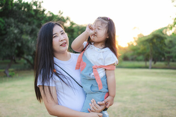 Fototapeta premium Happy Asian mother carrying her little daughter in the public park during golden hour sunset,A beautiful young Asian mother holding her cute little daughter in her arms while walking in the green park