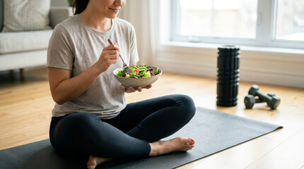 Happy young woman eating healthy green vegetable salad smiling sitting on yoga mat in living room after fitness workout session enjoying natural organic diet and peaceful active lifestyle