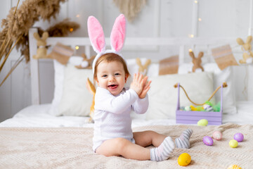 Fototapeta premium cute happy baby girl in bunny ears on the bed at home with a rabbit and painted eggs, easter concept, funny Easter baby is waiting for the holiday and rejoicing