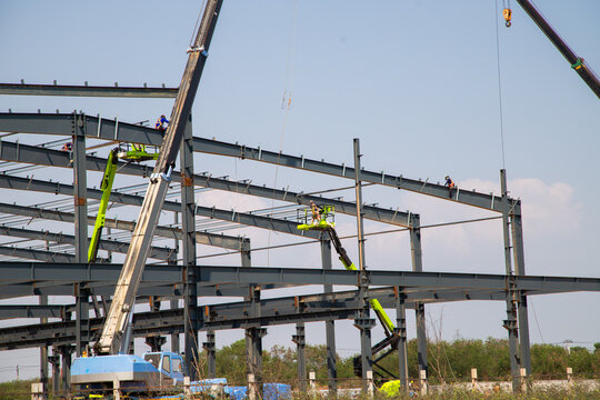 Large Steel-Frame Warehouse Structure Under Construction with Crane and Worker,Construction Workers on Cherry Picker Positioning High-Beams for New Facility