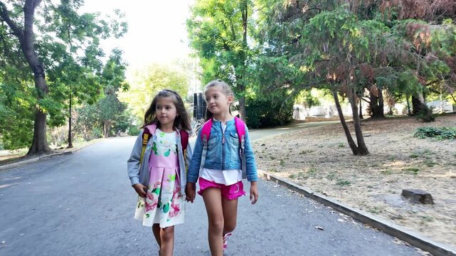 Happy children. Two girls with school backpacks walking in the park. Two little sisters walking together, talking. Best friends