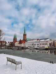 Obraz premium Frozen Trave river with snowy bench and Luebeck Old Town skyline in distance