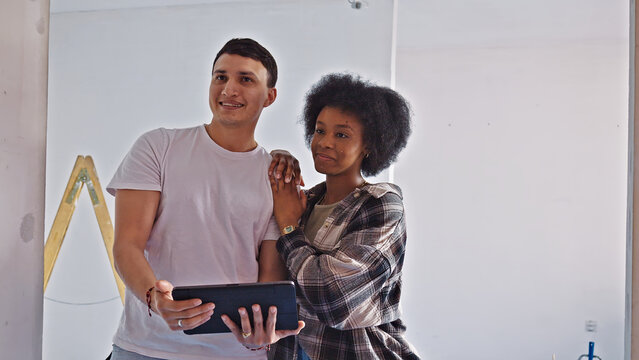 A young couple collaborates on a tablet in a modern, minimalist home under renovation. The man, in a white t-shirt, and the woman, in a plaid shirt, stand close.