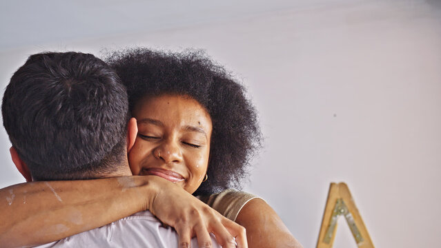 A couple embraces in a cozy home interior. The woman, with a relaxed expression, and the man, whose back is turned, seem to share a bond over their shared home improvement project.