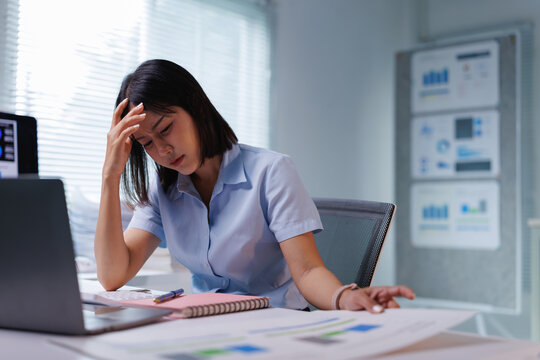 Young Asian woman at office desk late at night holding her head, exhausted and overwhelmed by workload, stress, and burnout from deadlines and paperwork