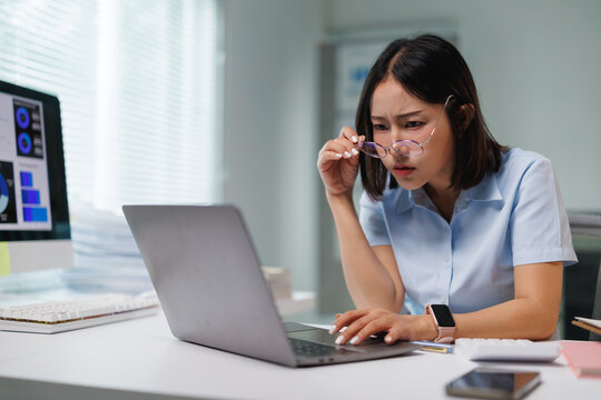Businesswoman experiencing poor vision and eye strain, holding up her glasses while concentrating on a laptop screen, dealing with frustration and confusion during work in an office