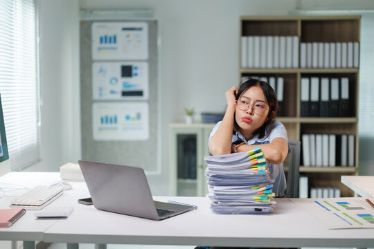 Young businesswoman feeling stressed and exhausted from a large stack of paperwork, looking bored and overwhelmed by her heavy workload in a modern office