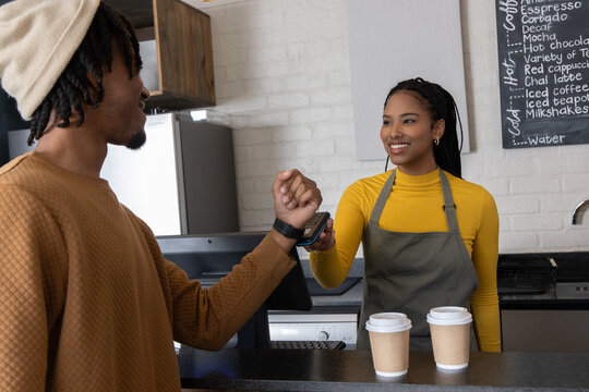 African American customer and barista tapping smartwatch at counter with card reader, olive apron
