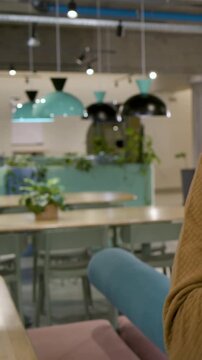 Vertical video: Fiddling man in sweater holding bottle, working at laptop in lounge with headphones