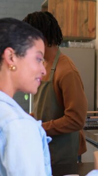 Vertical video: Smiling woman in denim with box paying at cafe till as staff using POS, copy space