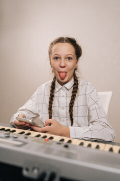 Cheerful young girl with braided pigtails sitting at electronic musical keyboard, using cellphone and playfully sticking out tongue, expressing fun and creativity during music learning session