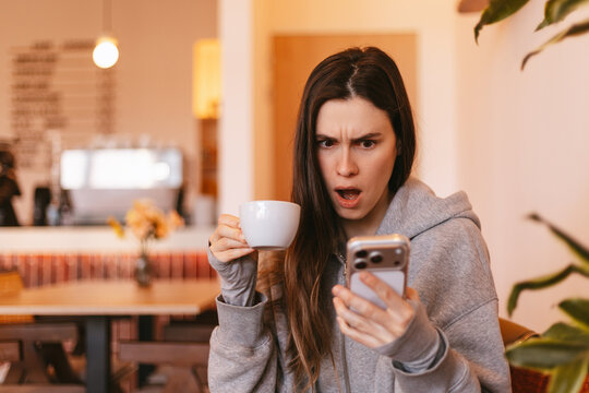 Shocked and outraged young woman in grey hoodie holding coffee cup and looking at smartphone with wide open mouth in cozy cafe interior during morning break time reading unbelievable news