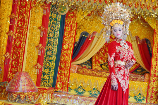A portrait of a Minangkabau bride in a detailed red and gold traditional dress and elaborate headdress, posed against an ornate wedding backdrop