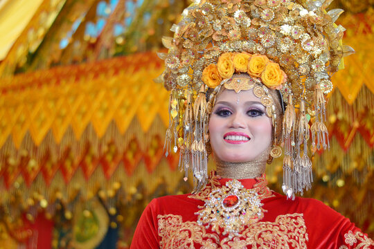 Close-up of a beautiful Acehnese bride wearing a majestic golden headdress and intricate red traditional wedding attire, smiling radiantly
