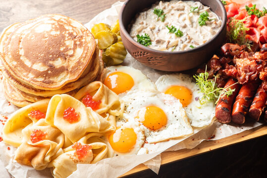 Traditional Russian breakfast board with pancakes, blini with red caviar, fried eggs, grilled sausages, bacon, pickles, and a bowl of creamy mushroom sauce on parchment paper.