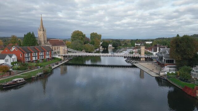A serene river flows past a town with a prominent church spire and a white suspension bridge. Boats are docked along the bank.