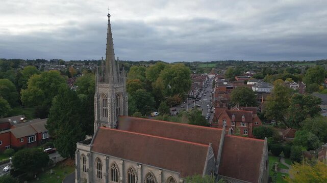 An aerial view shows a church with a tall spire, surrounded by autumnal trees and a quaint village street filled with cars.