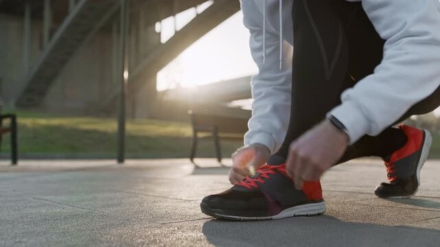 Sportsman in athletic clothing fastening his running shoes beneath a bridge at sunrise. The image captures preparation for a morning run, emphasizing urban fitness, motivation, and active lifestyle.