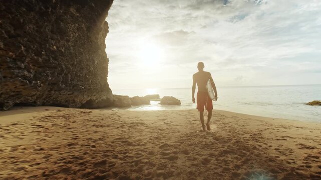 Surfer walks out of a coastal cave at sunset, carrying a surfboard along an empty beach. Warm golden sunlight illuminates the scene, casting long shadows and creating a serene, tranquil atmosphere