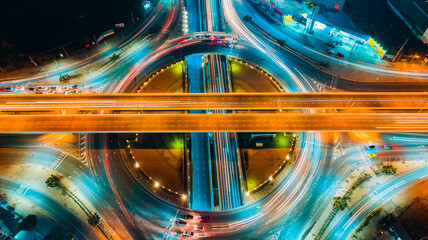 Aerial top view of circular highway interchange at night, glowing orange light trails crossing central overpass ring, captured with high angle and neon tones, symbolizing fast transportation, © AU USAnakul