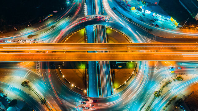 Aerial top view of circular highway interchange at night, glowing orange light trails crossing central overpass ring, captured with high angle and neon tones, symbolizing fast transportation,
