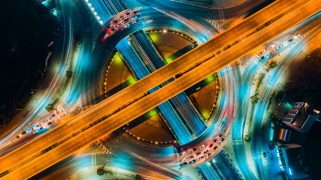 Night drone shot of multilayer highway junction with circular core and diagonal overpass, long exposure light trails forming dynamic geometry, wide-angle perspective emphasizing motion,