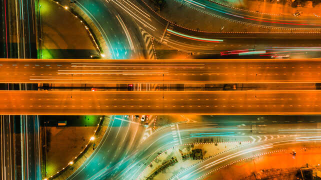 Top view aerial shot of symmetrical highway interchange at night, glowing orange traffic streams crossing smooth curved roads, captured with high angle and neon lighting, representing modern transport