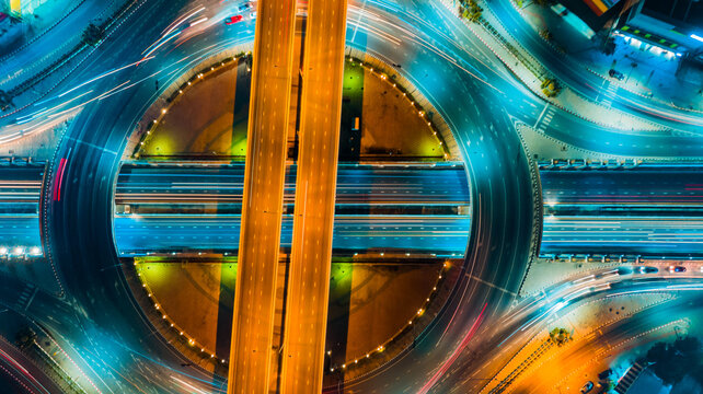 Aerial top view of diagonal highway crossing circular interchange at night, bright orange light trails slicing through glowing blue roads, captured with high angle and neon lighting,