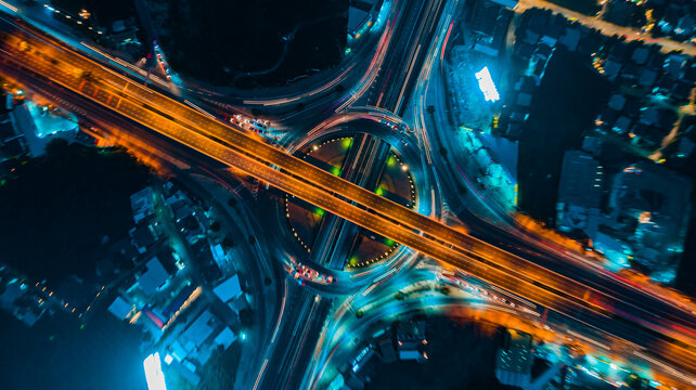 Top view aerial shot of symmetrical highway interchange at night, glowing orange traffic streams crossing smooth curved roads, captured with high angle and neon lighting,