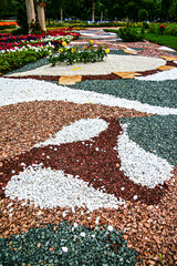 Fototapeta premium Artistic garden pathway made of colorful pebbles forming abstract patterns, surrounded by flowers and greenery in a landscaped park setting.