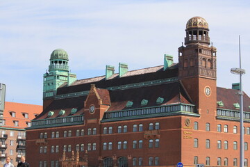 Obraz premium Historic red‑brick Malmö Central Station building with arched windows and tiled roof on a sunny day, cars and pedestrians in front and Central Post Office clocktower in background, Sweden.