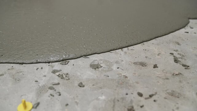 A close-up video shows a self-leveling compound being poured onto a concrete subfloor. The wet grey cement mixture slowly spreads and covers the uneven surface, preparing it for new flooring.