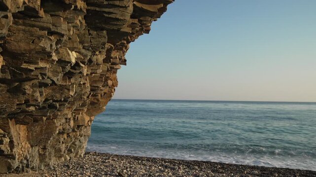 Ocean cliff beach view of gentle waves rolling onto a pebble shore beneath a rugged stone precipice at sunrise