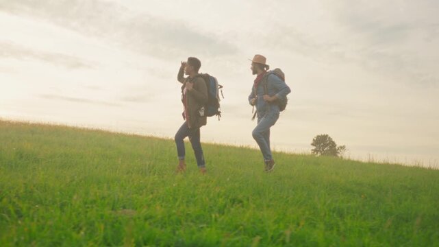 Rolling hill trek with two hikers stride toward horizon soft sunrise glow backpacks denim hat open grassland calm