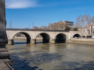 Historical stone bridge with multiple arches spanning across the Mtkvari River in Tbilisi, Georgia, with city buildings and bare trees in the background under a clear blue sky. © Microscope